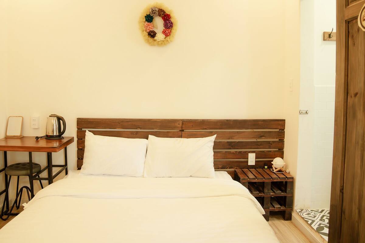 A neatly arranged bedroom with a wooden headboard, a small bedside table with a kettle, a nightstand with a lamp, and a decorative wall hanging.