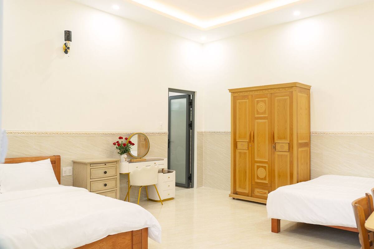 A hotel room with two beds, a wooden wardrobe, a vanity with a mirror, and a small table with a vase of flowers.