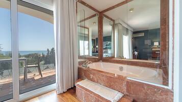 A modern bathroom with a large marble bathtub, a glass-enclosed shower, and a balcony with a view of the sea.