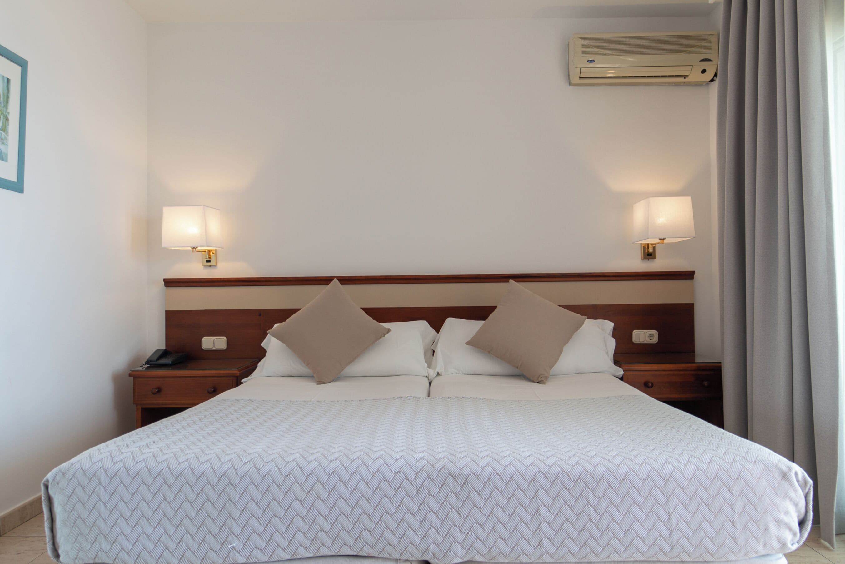 A hotel room with a large bed, two bedside tables, a wall-mounted air conditioner, and two wall lamps.