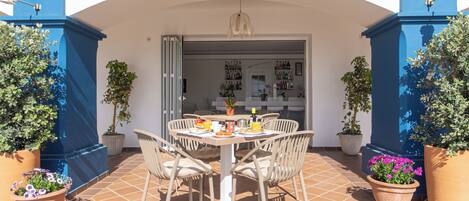 Une terrasse avec une table ronde et des chaises, entourée de plantes en pot et dotée d’un sol carrelé.