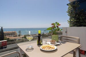 Une table avec un verre de vin blanc, une bouteille d’huile d’olive, une assiette de collations et une plante en pot, le tout sur un balcon donnant sur la mer.