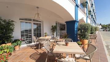 Un patio al aire libre con una mesa y sillas, rodeado de plantas en macetas y un edificio con detalles azules.