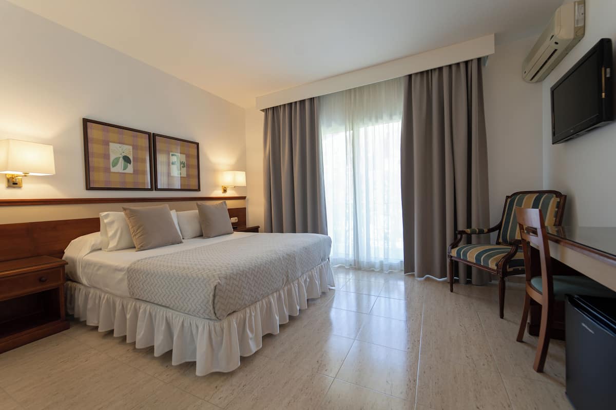 Habitación de hotel con cama, dos mesitas de noche con lámparas, una silla, televisor y un amplio ventanal con cortinas.