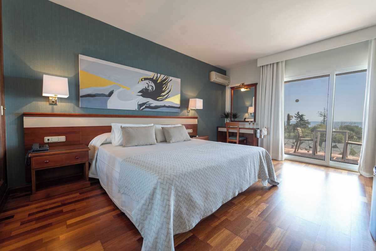 Habitación de hotel con una cama grande, dos mesitas de noche, un aparador, un cuadro en la pared y un balcón con vistas.