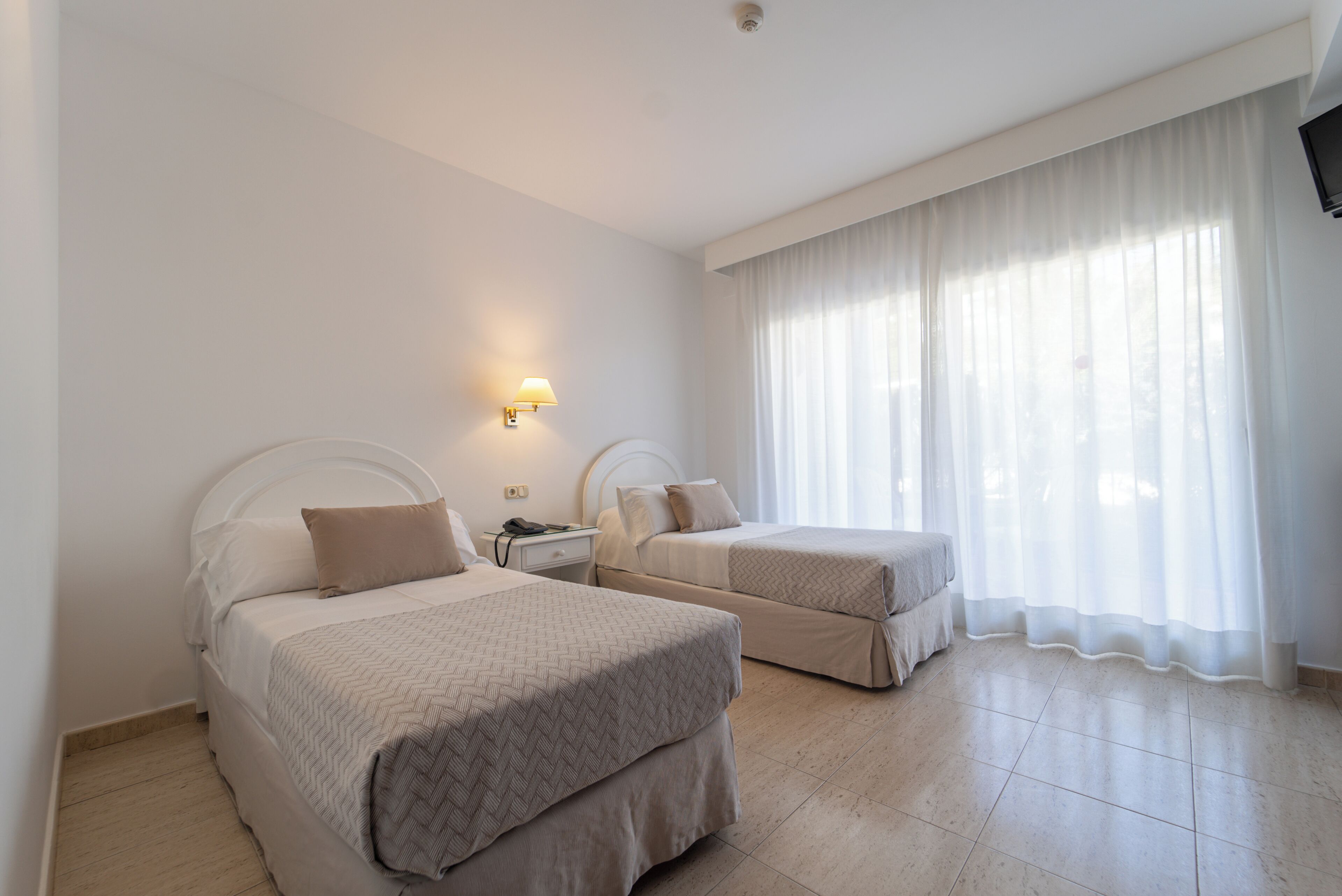 A hotel room with two beds, a nightstand, a lamp, and a large window with sheer curtains.