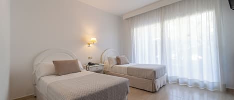A hotel room with two beds, a nightstand, a lamp, and a large window with sheer curtains.