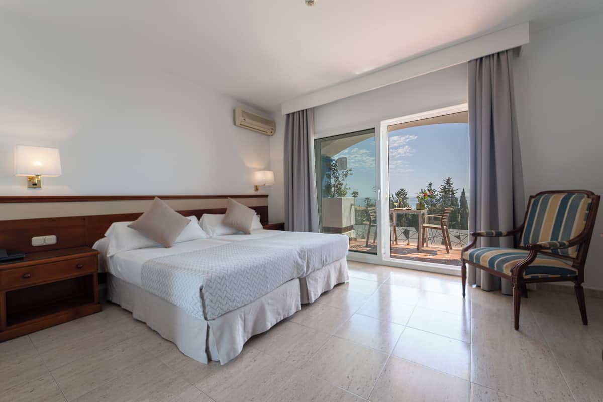 Una habitación de hotel con cama, mesita de noche, silla, balcón con mesa y sillas, y vista a árboles y edificios.