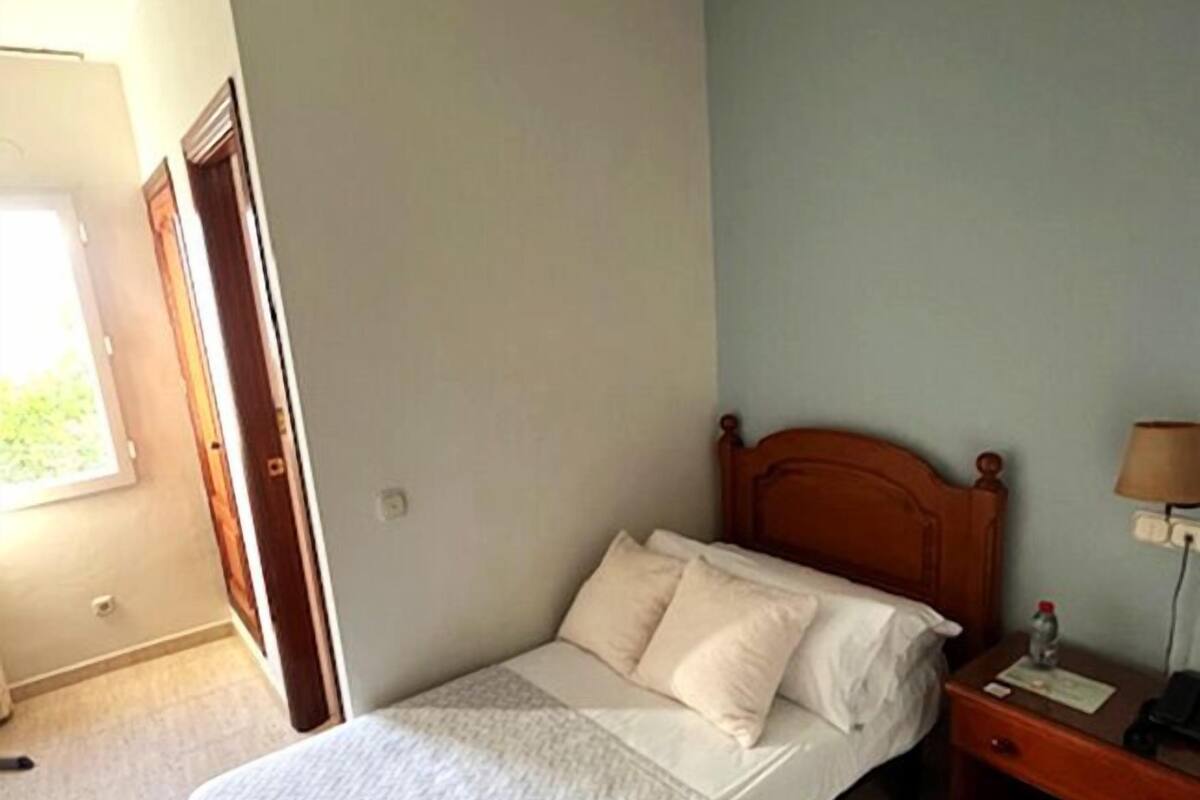 Una habitación pequeña con cama individual, cabecera de madera, mesita de noche con lámpara y ventana que deja entrar luz natural.