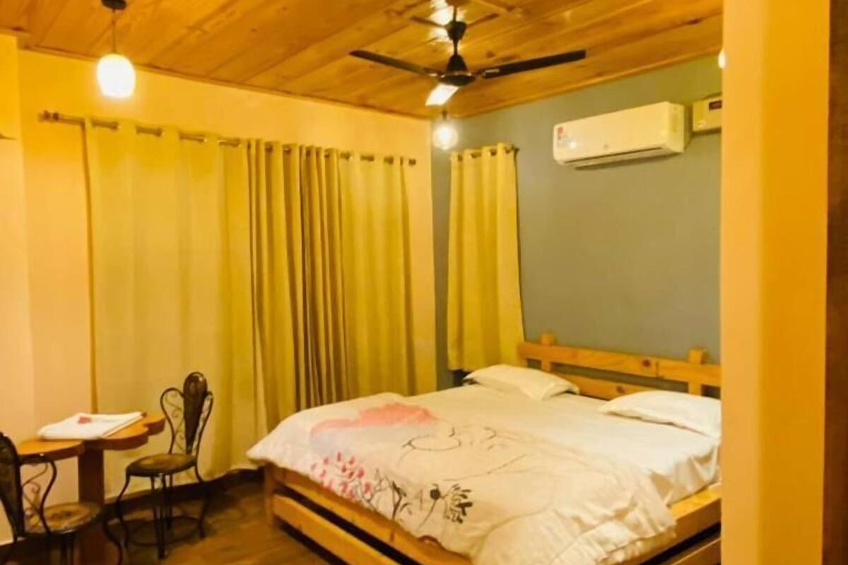 A room with a wooden ceiling, a bed with a floral bedspread, a desk with a chair, and an air conditioning unit on the wall.