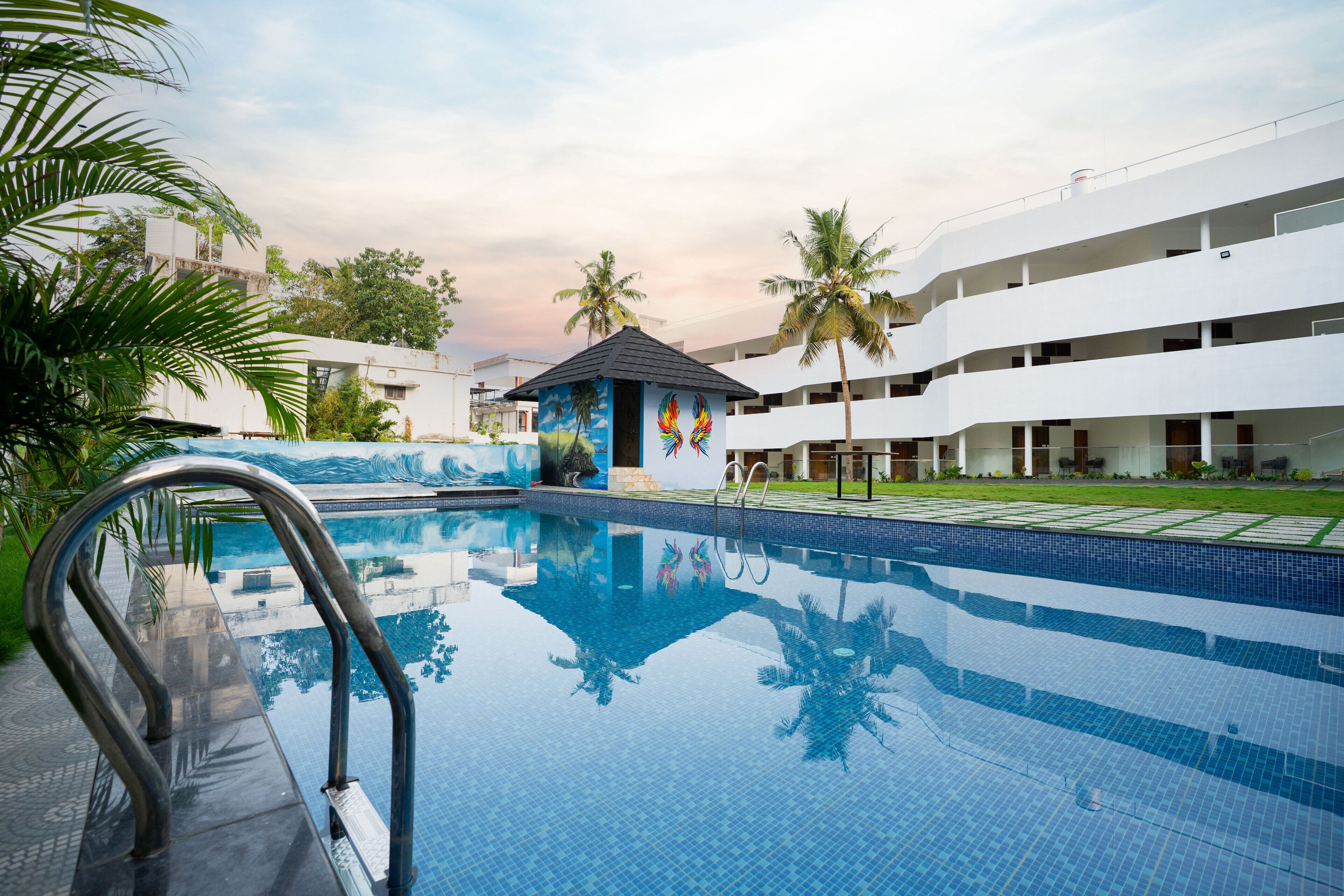 Piscina con escalera metálica, rodeada de palmeras y un edificio con un mural.