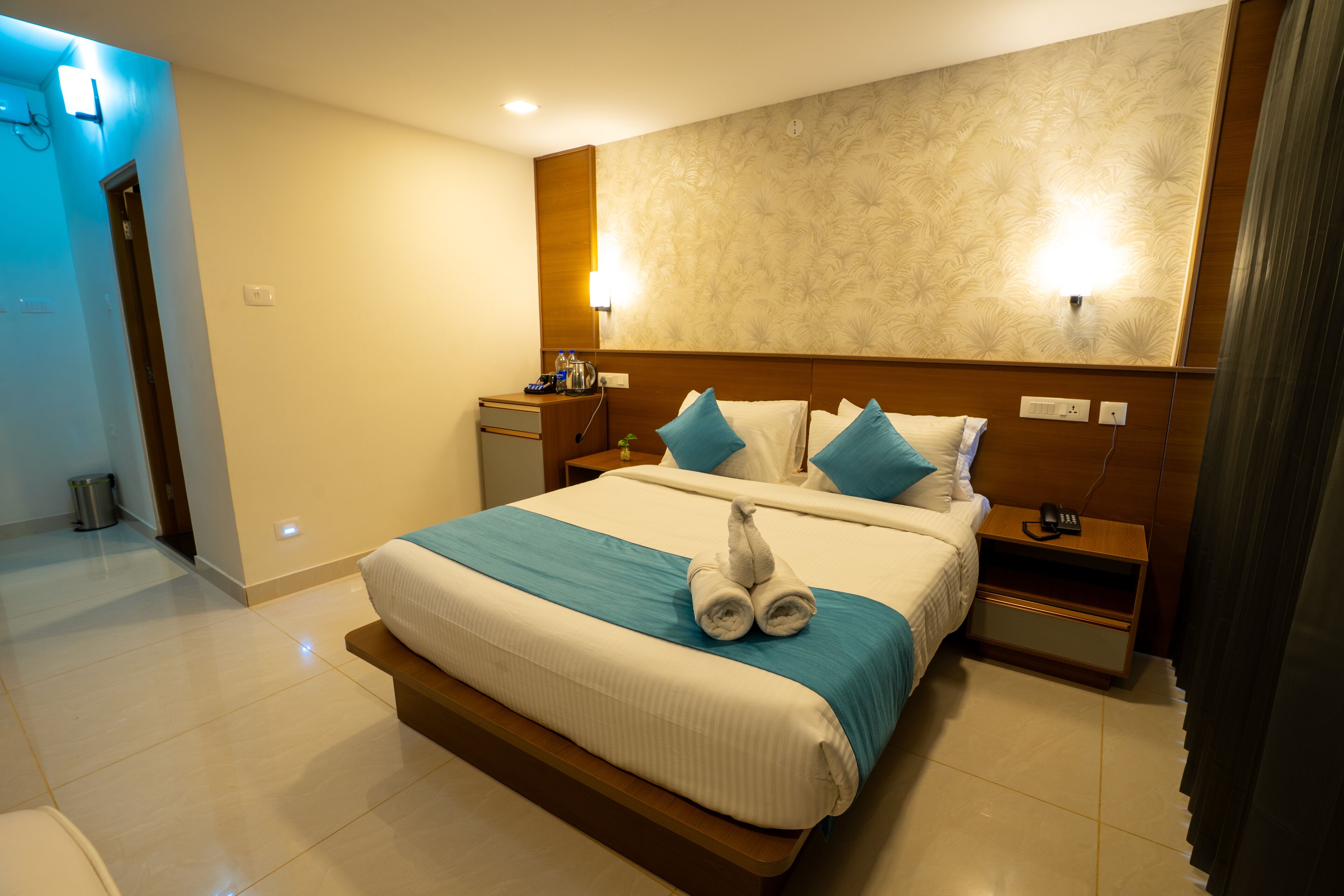 A hotel room with a large bed, a nightstand, a wall-mounted lamp, and a bathroom visible in the background.