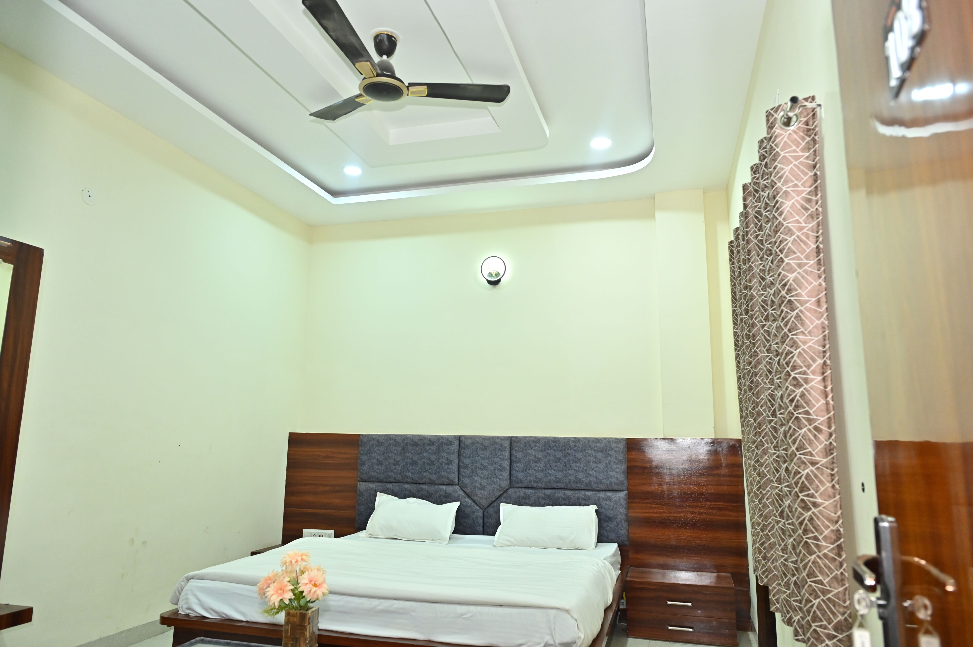 A hotel room with a bed, a ceiling fan, a security camera, and a patterned curtain.
