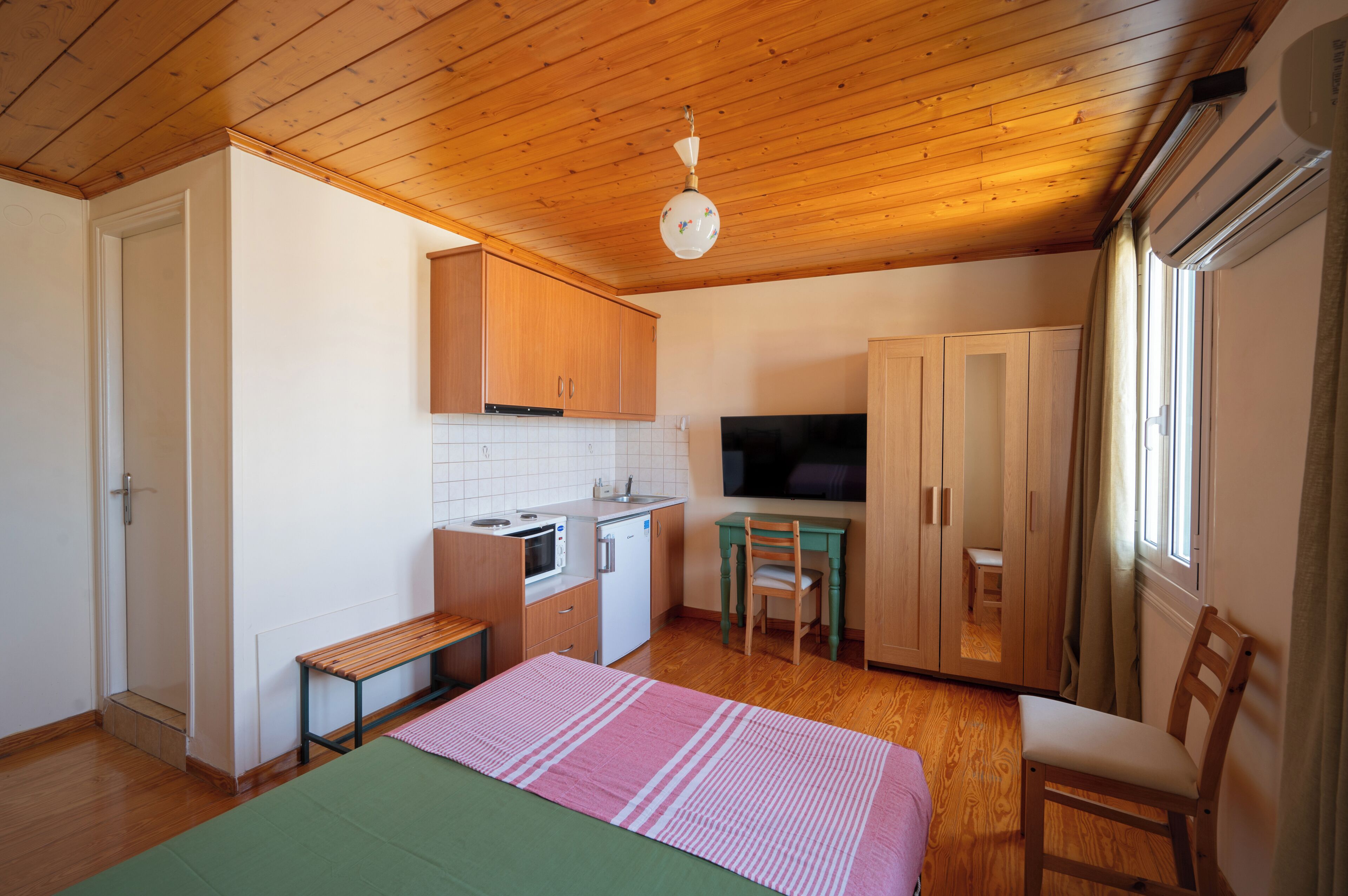A compact kitchen with wooden cabinets, a microwave, and a sink. There is a dining area with a table and chairs, and a bed with a green and pink striped blanket.