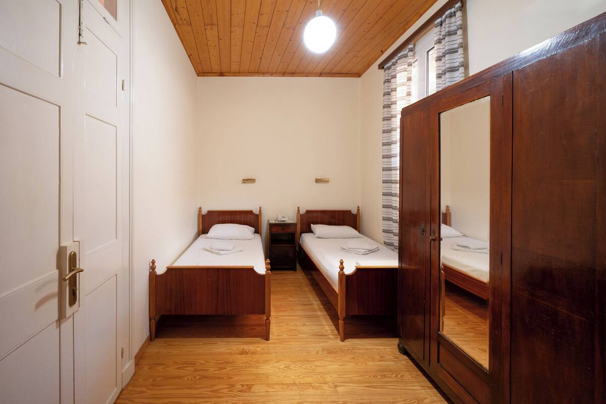 A room with two wooden beds, a wardrobe, and a small table with a lamp.