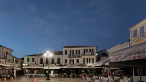 An outdoor café area with tables and chairs, surrounded by buildings with various signs and awnings. - Hotel Patrai (Lefkada Town)