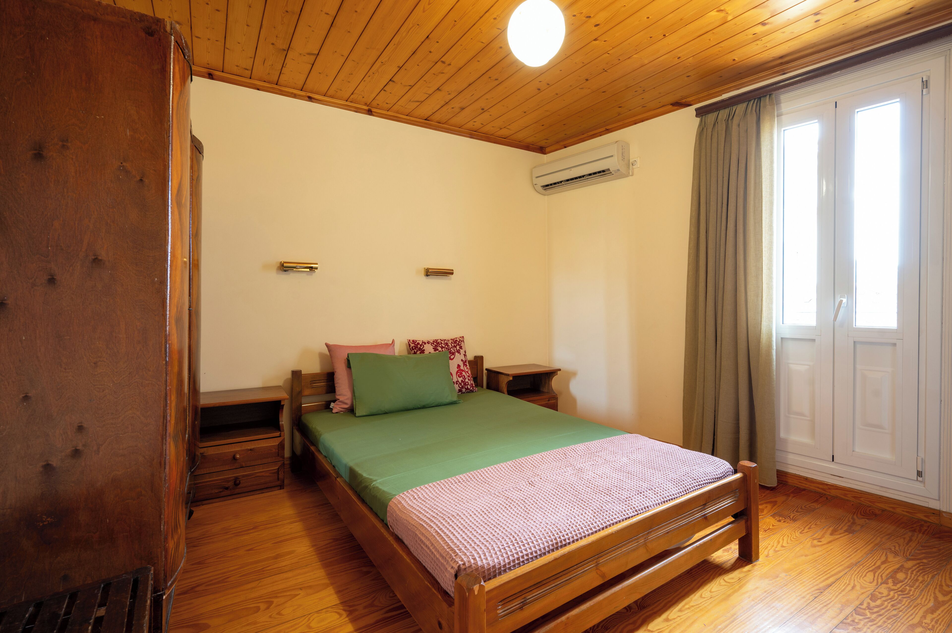 Una cama con estructura de madera, cubierta con edredón verde y rosa, una mesita de noche de madera, y una ventana con cortinas.