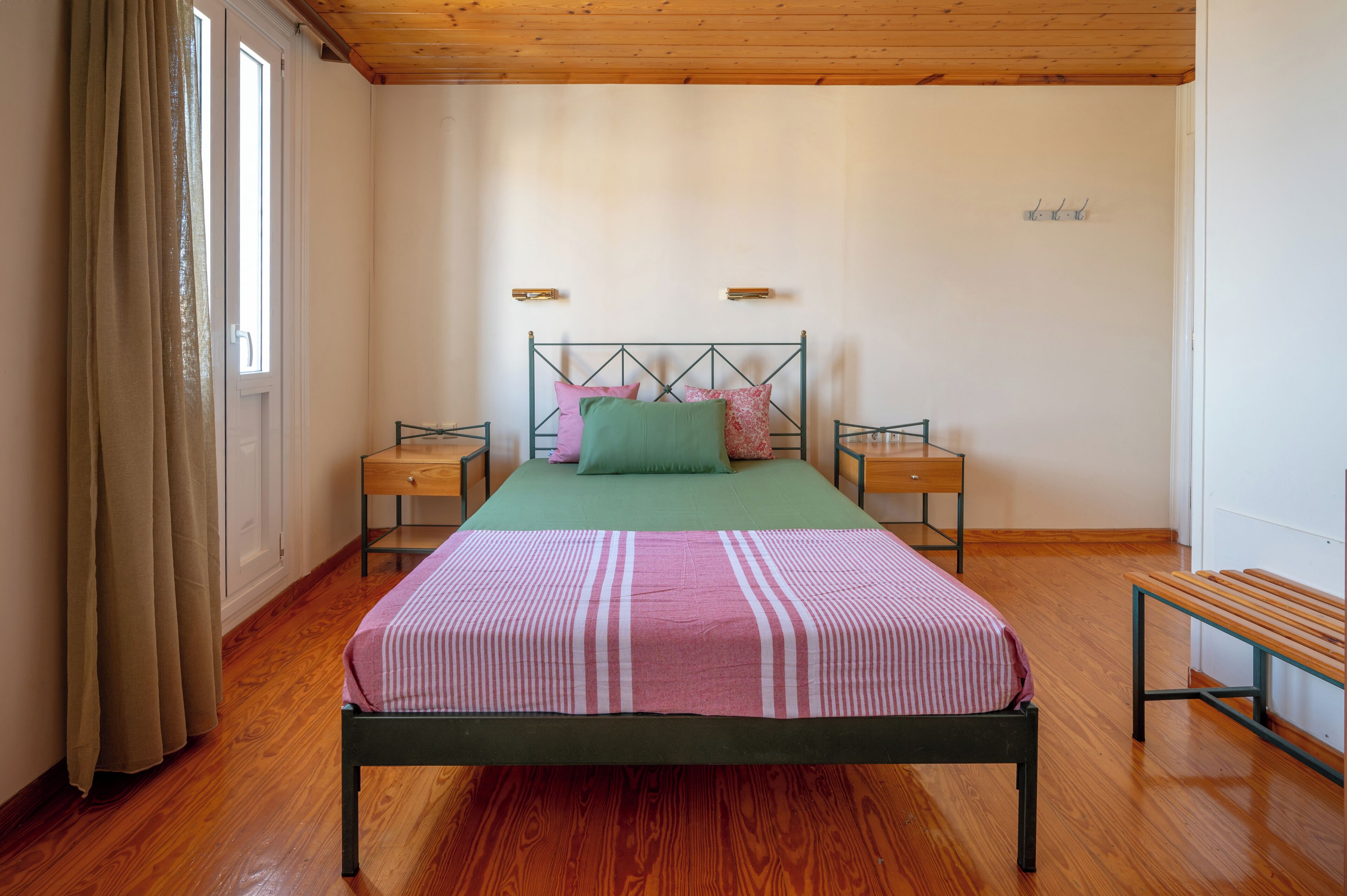 Un dormitorio con cama, dos mesitas de noche, un banco y una ventana con cortinas.