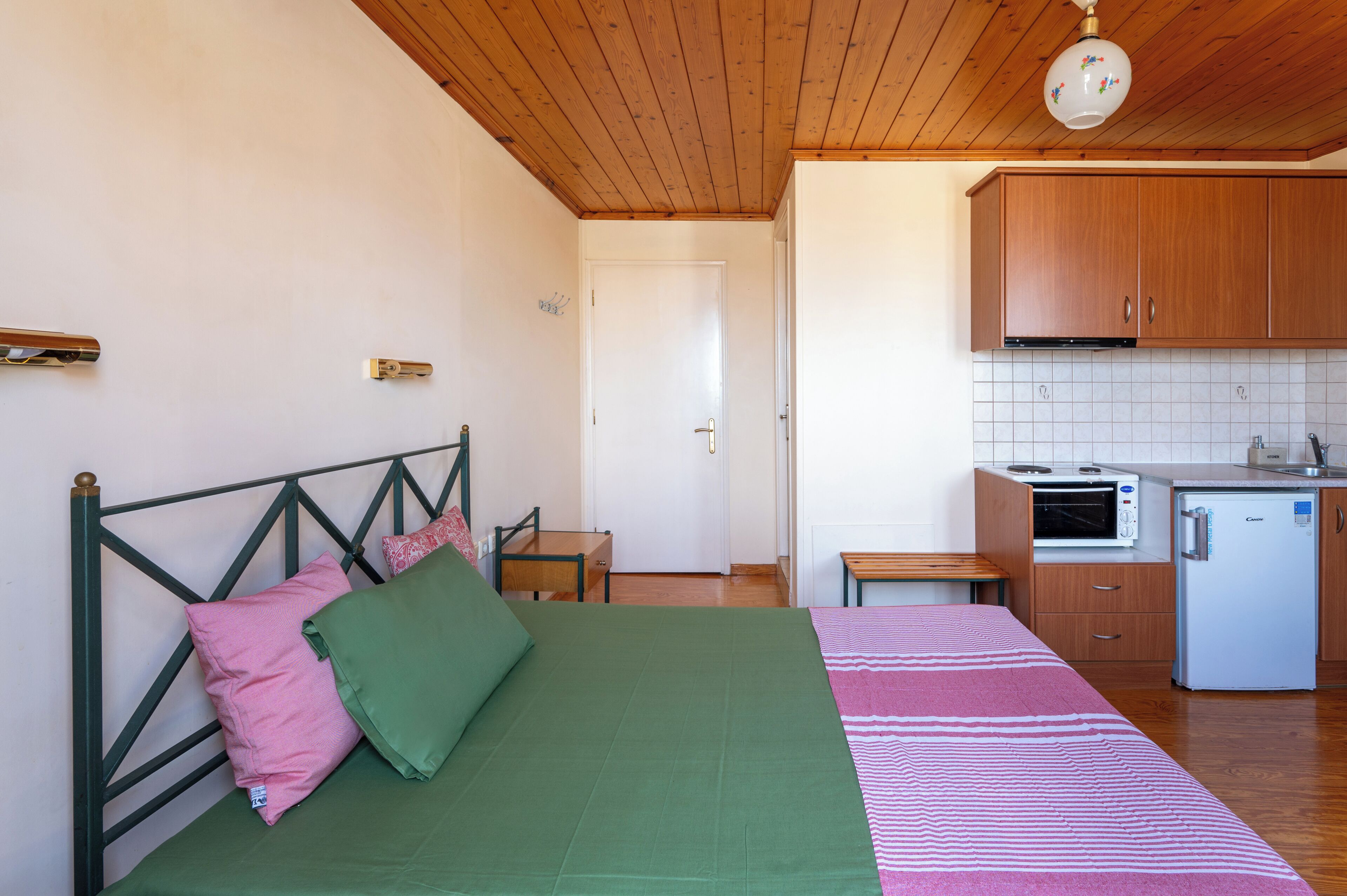 Ein Schlafzimmer mit Bett, Kissen, einem Nachttisch und einem kleinen Holzsessel. Angrenzend an das Schlafzimmer befindet sich eine Küchenzeile mit einer Mikrowelle, einem Kühlschrank und Holzschränken.