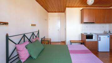 Ein Schlafzimmer mit Bett, Kissen, einem Nachttisch und einem kleinen Holzsessel. Angrenzend an das Schlafzimmer befindet sich eine Küchenzeile mit einer Mikrowelle, einem Kühlschrank und Holzschränken.