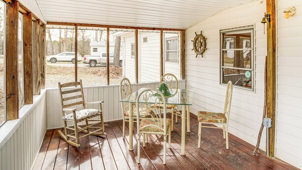 En inglasad veranda med ett bord och stolar, en gungstol och utsikt över ett vitt hus och parkerade fordon.