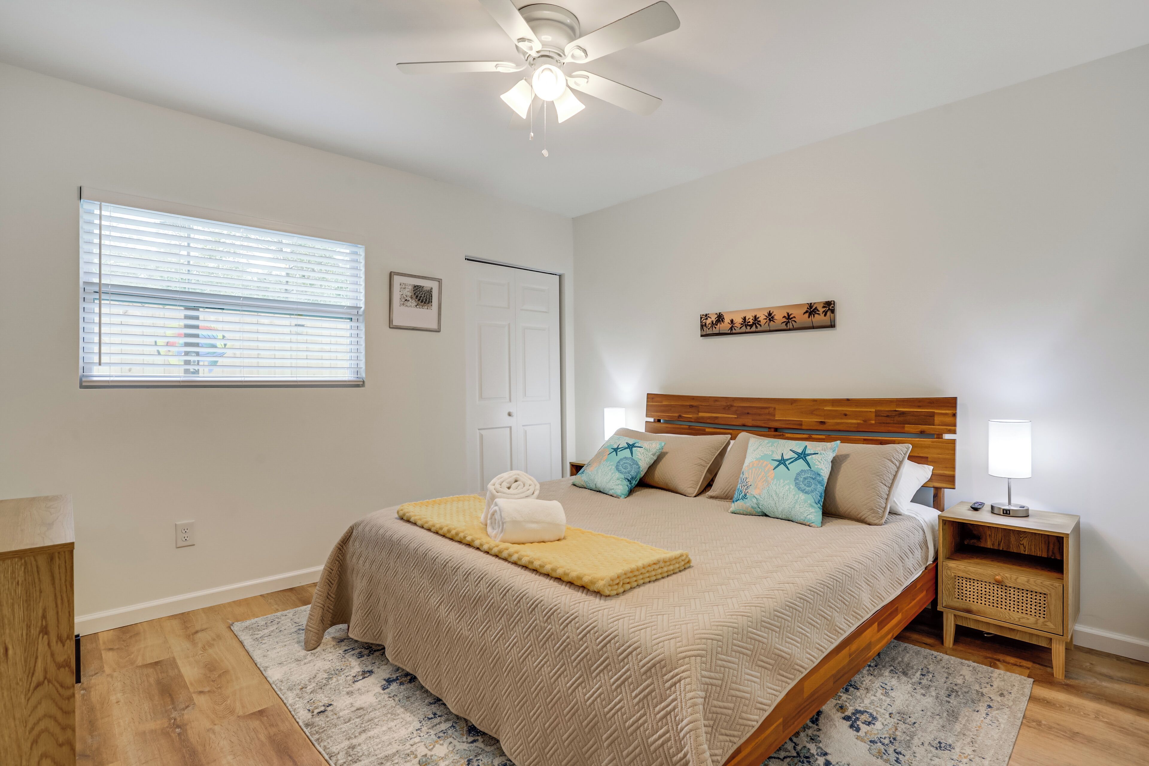 A bedroom with a bed, a ceiling fan, a nightstand, a window with blinds, and a door.