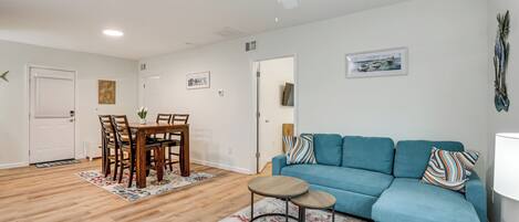 A living room with a blue sofa, a dining table with chairs, a ceiling fan, and a door.