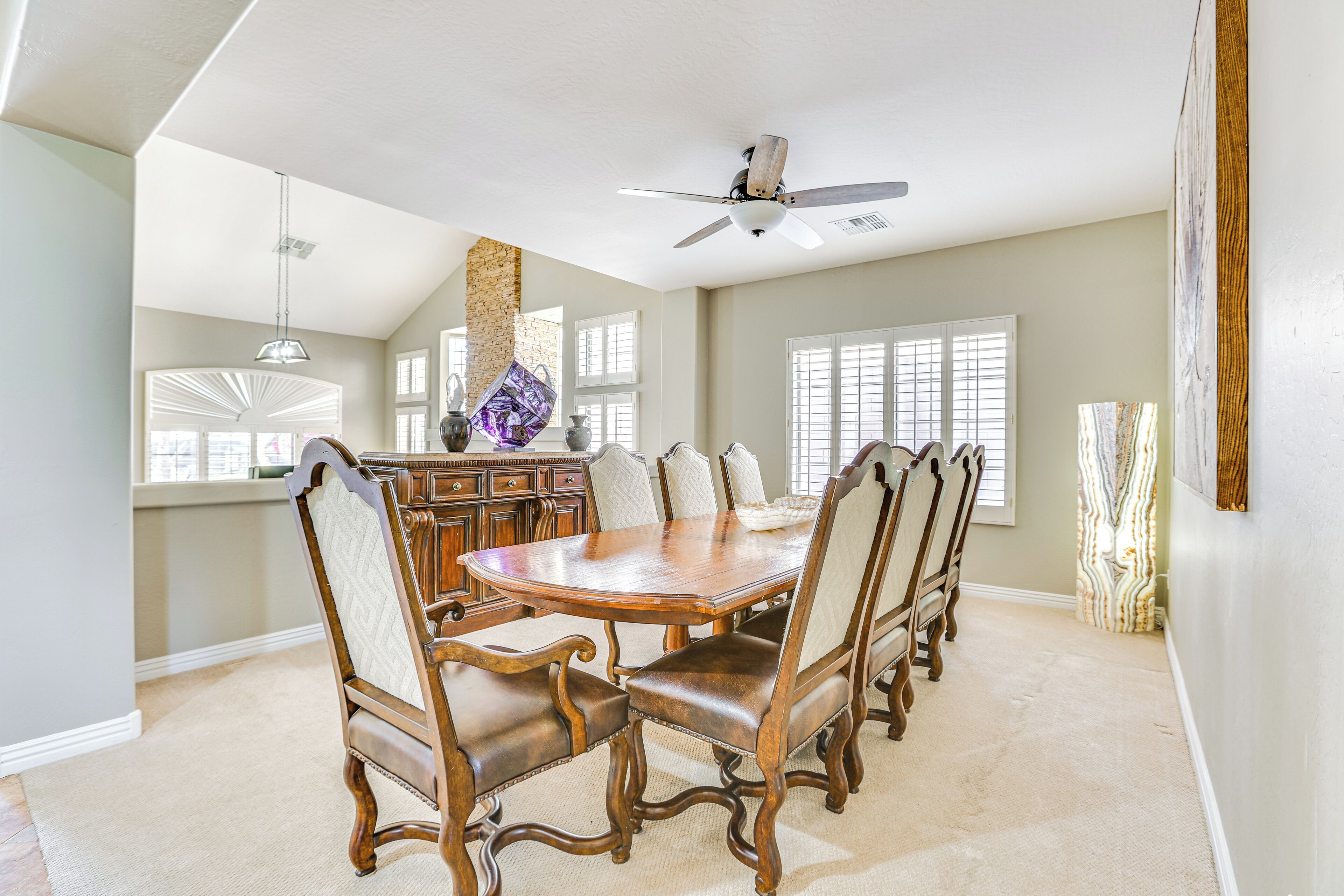 A dining room with a large wooden table and chairs, a ceiling fan, and a view of the kitchen area.