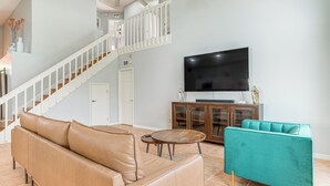 Ein Wohnzimmer mit einem beigefarbenen Ledersofa, einem türkisfarbenen Sessel, einem Holztisch, einem Fernseher und einer Treppe.