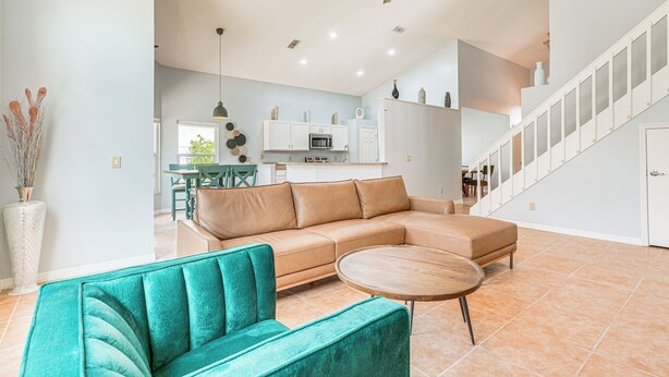 Ein großzügiger Wohnbereich mit einem türkisfarbenen Sofa, einem Holztisch und einem beigefarbenen Ecksofa. Eine Deckenleuchte und Einbaustrahler sind zu sehen.