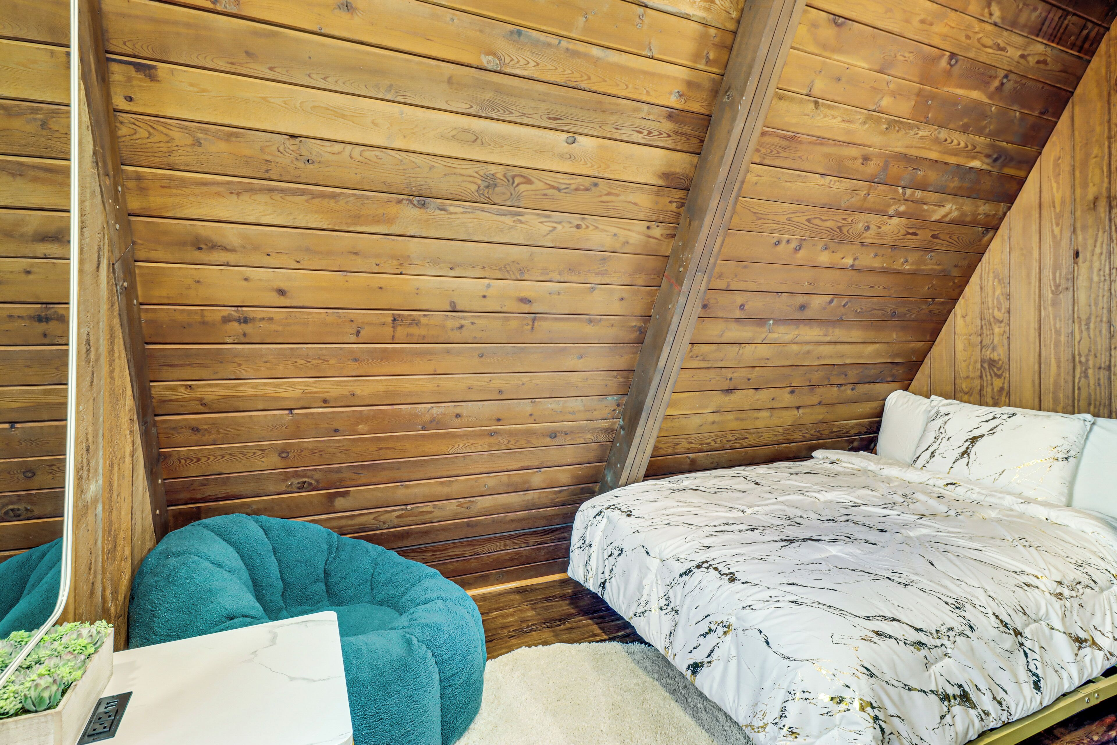 Une chambre mansardée chaleureuse avec des lambris en bois au plafond, un lit recouvert d’une couvre-lit à motifs, un fauteuil en forme de poire de couleur turquoise et une petite plante posée sur une table de chevet.