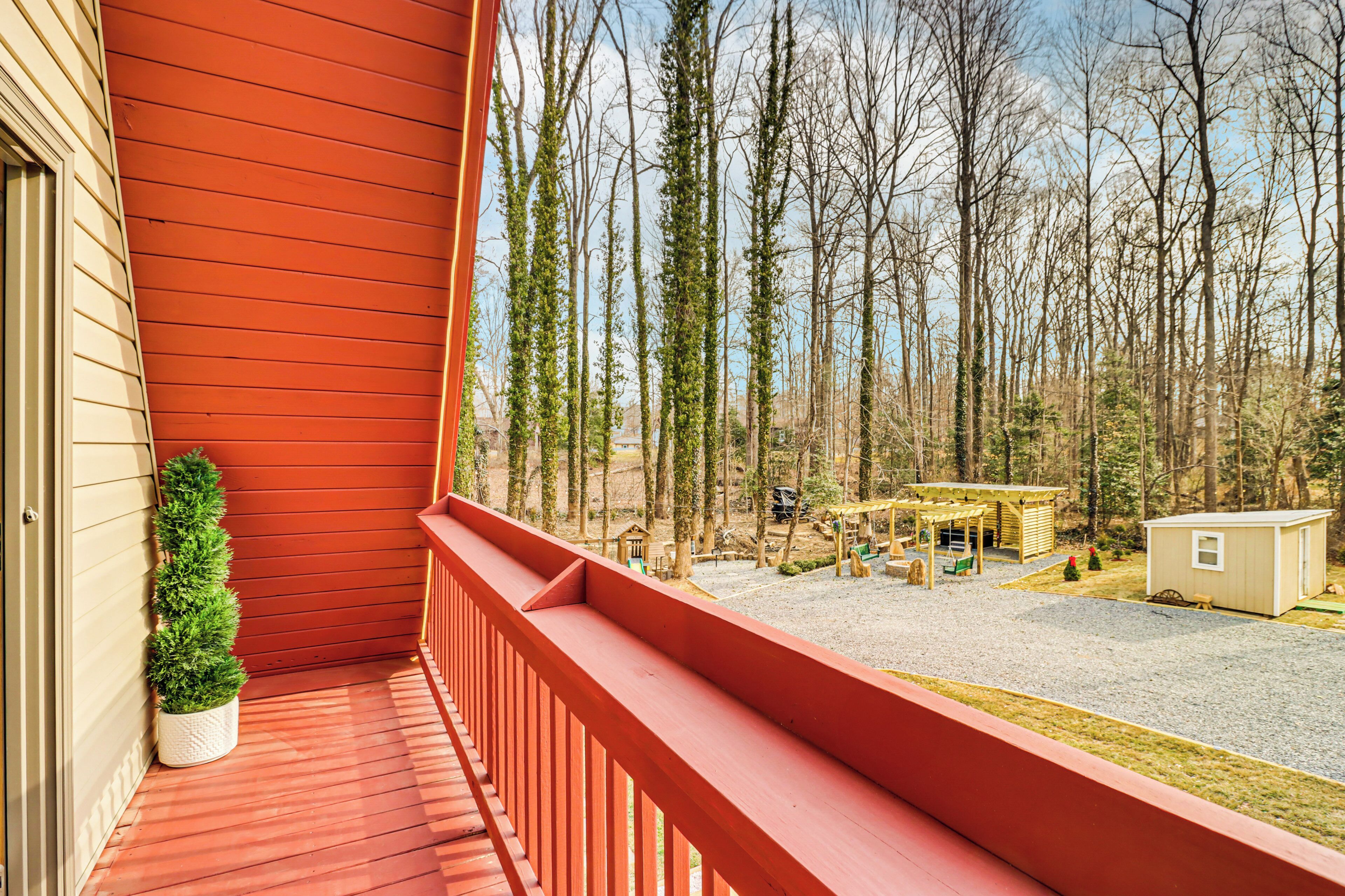 Une terrasse en bois avec une rambarde rouge, une petite plante en pot et une vue sur une zone boisée agrémentée d’un belvédère et d’un abri de jardin.