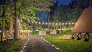 Een pad dat leidt naar een rustieke lodge, omringd door bomen, met lichtjes en een speeltuintje.