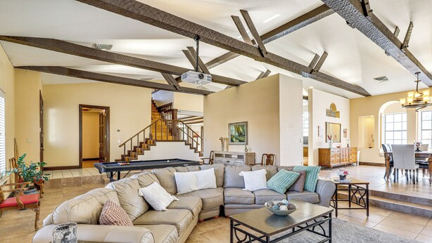 A spacious living room with a sectional sofa, a coffee table, and a pool table. There is a staircase leading to an upper level.
