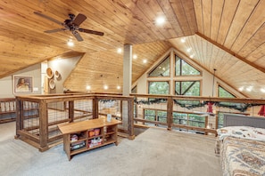 A spacious loft with a high ceiling, wooden beams, a ceiling fan, and a large window offering a view of the outdoors.