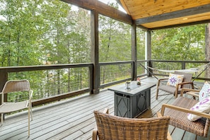 A covered outdoor deck with wicker furniture, a grill, and a view of the surrounding forest.