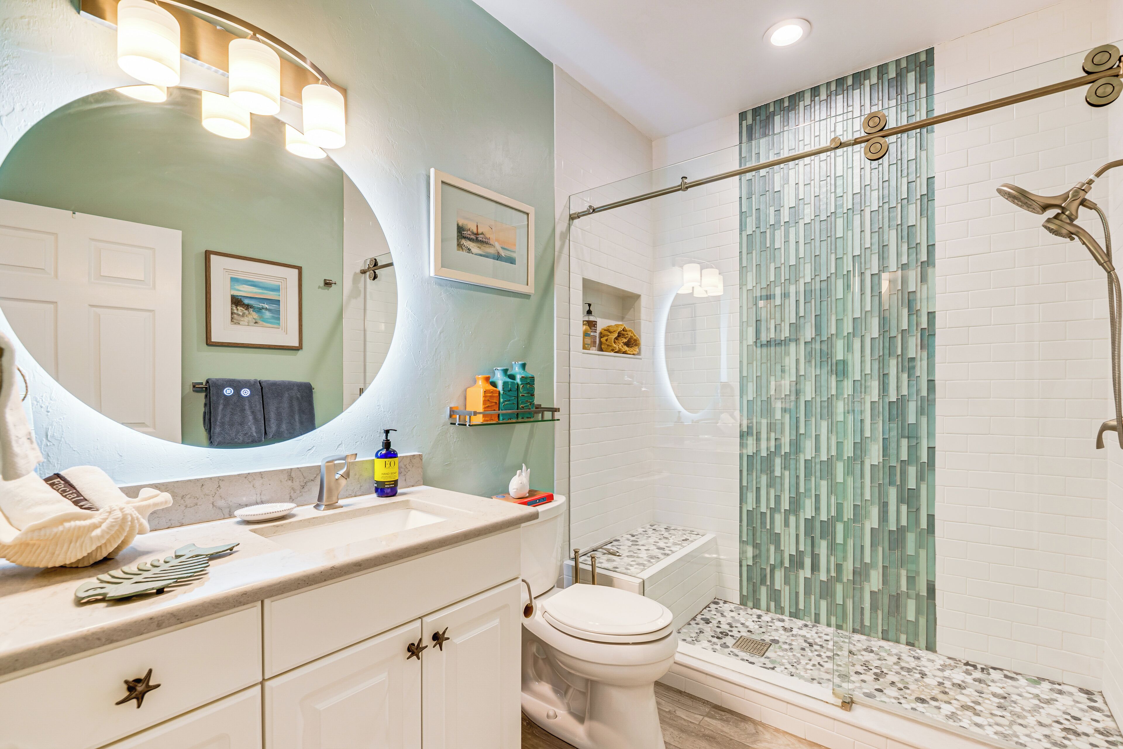 Un baño con tocador blanco, un espejo grande, inodoro, ducha con puerta de cristal y pared con azulejos.