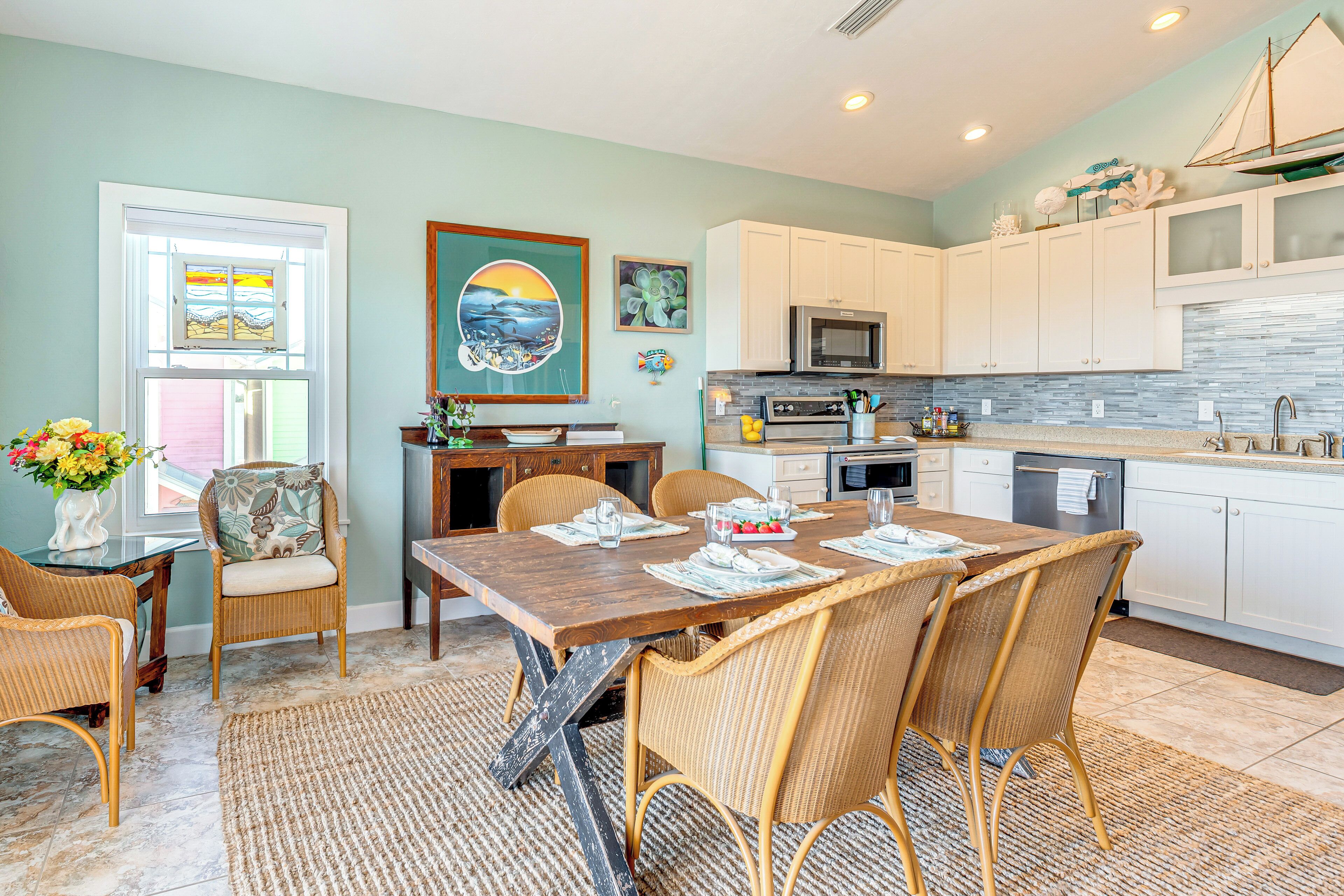 A dining area with a wooden table and wicker chairs, a kitchen with white cabinets and a microwave, and artwork on the walls.