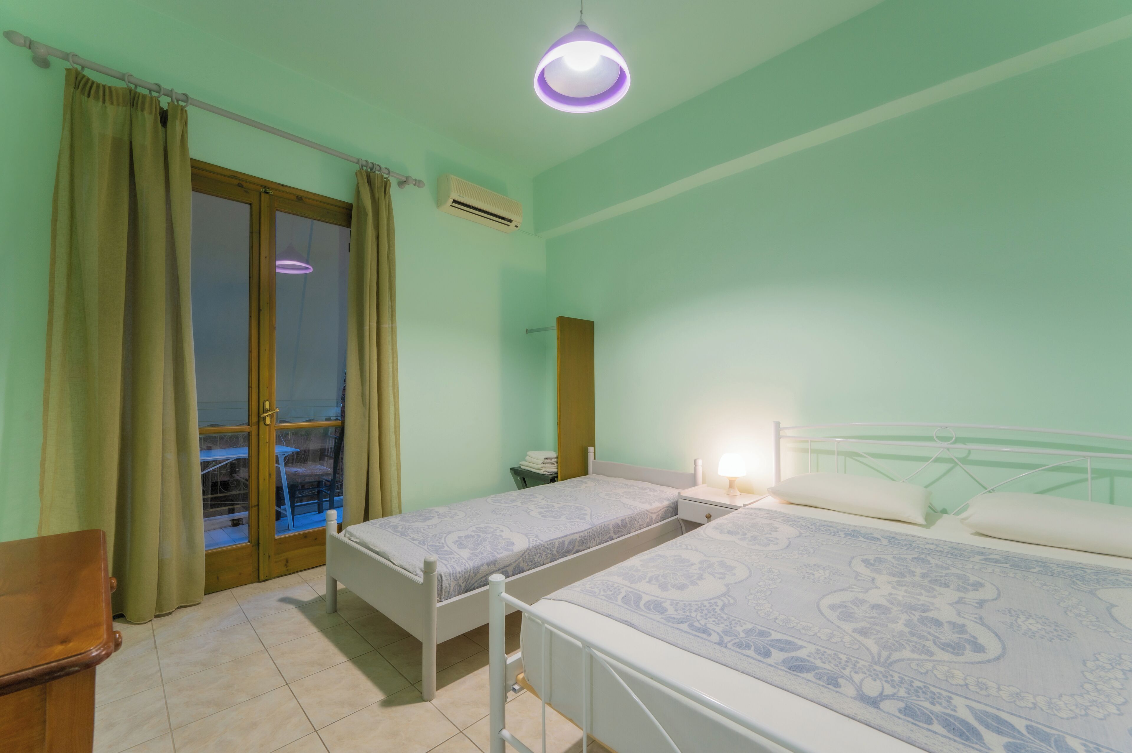 A room with two single beds, a wooden chair, a desk, a window with curtains, and a ceiling light.