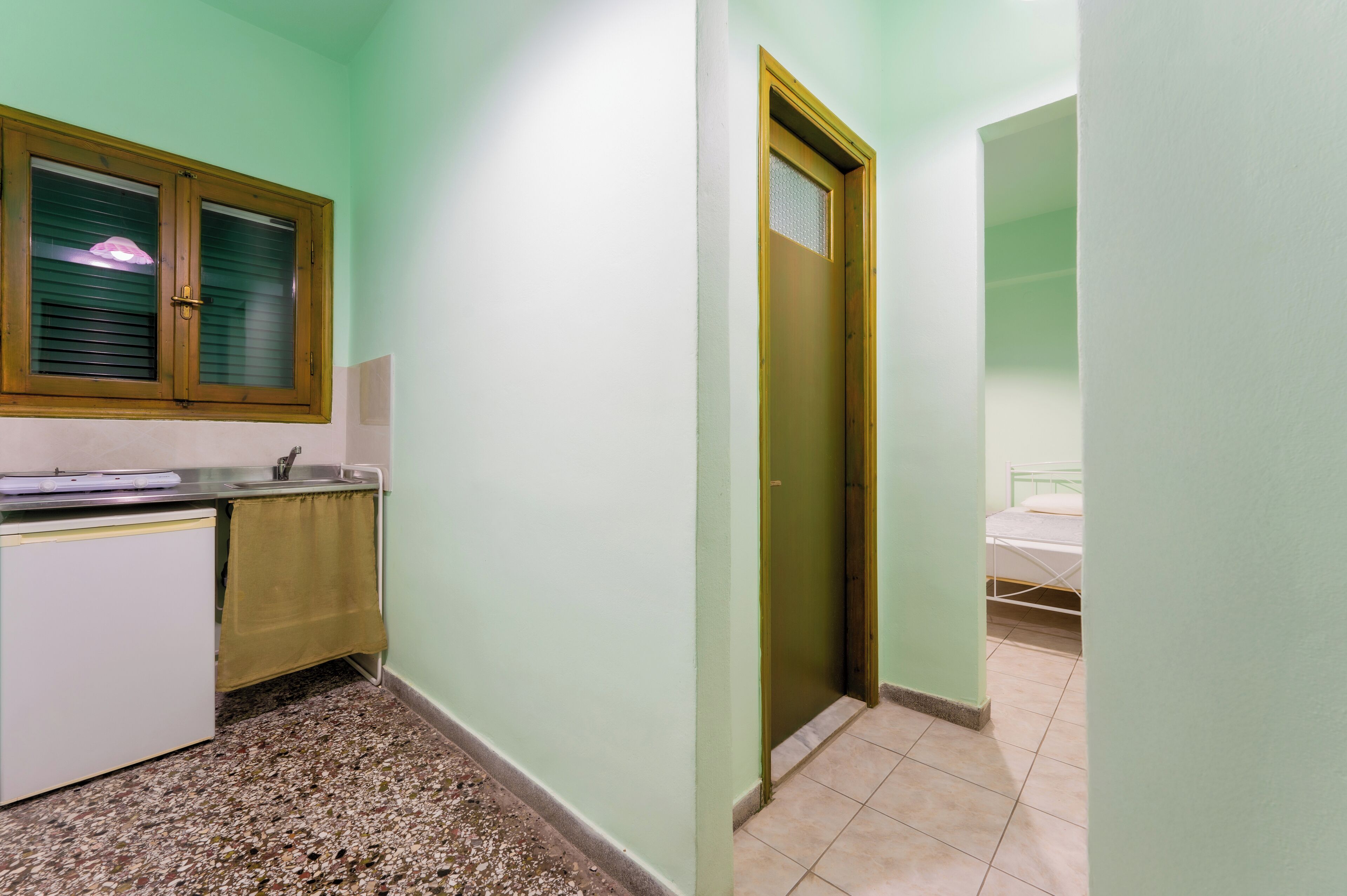 A narrow hallway with a kitchenette on the left, a door on the right, and a bed in the adjacent room.