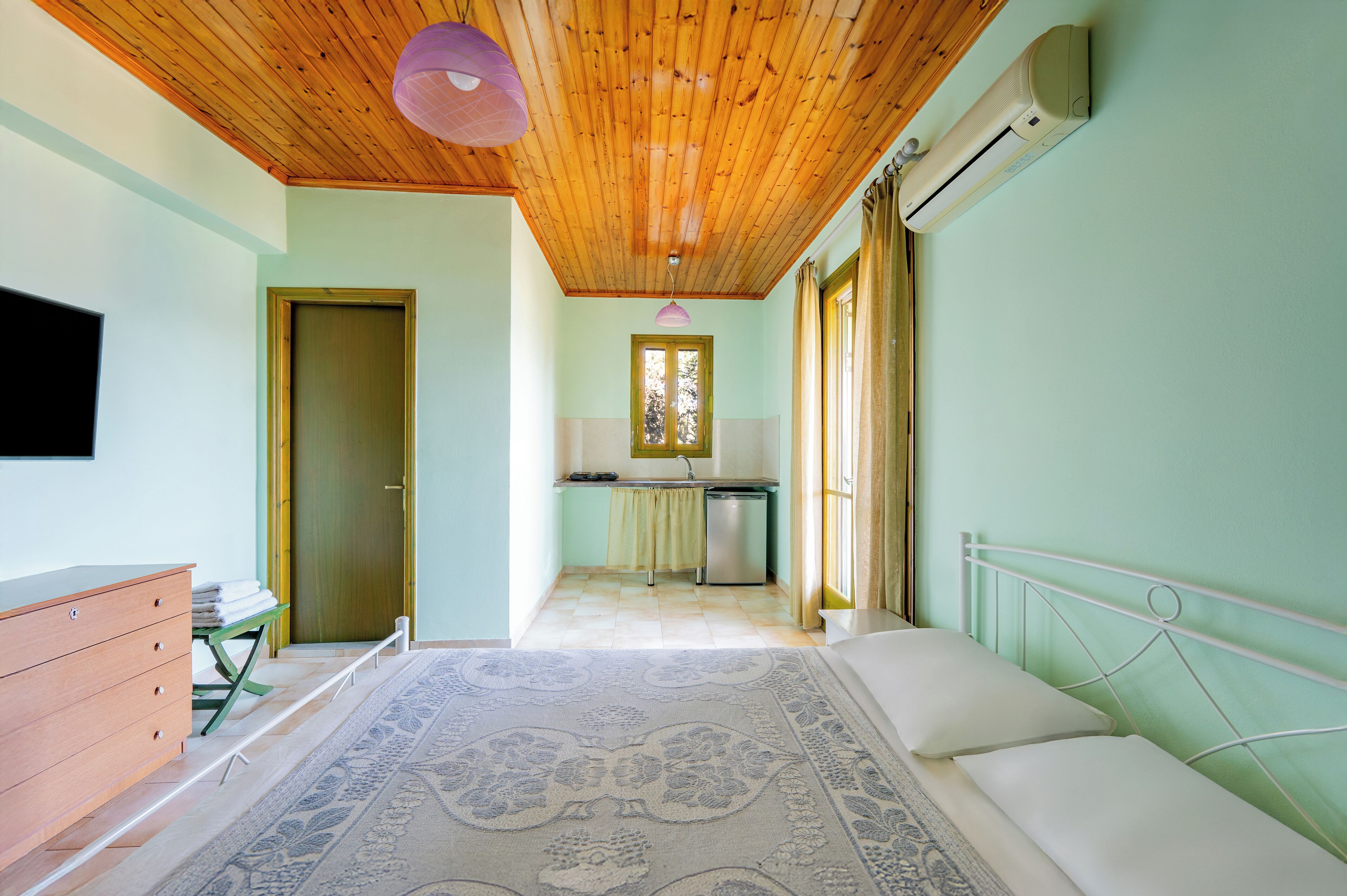 Un dormitorio con techo de madera, cama, cómoda, pequeña cocina americana y ventana.