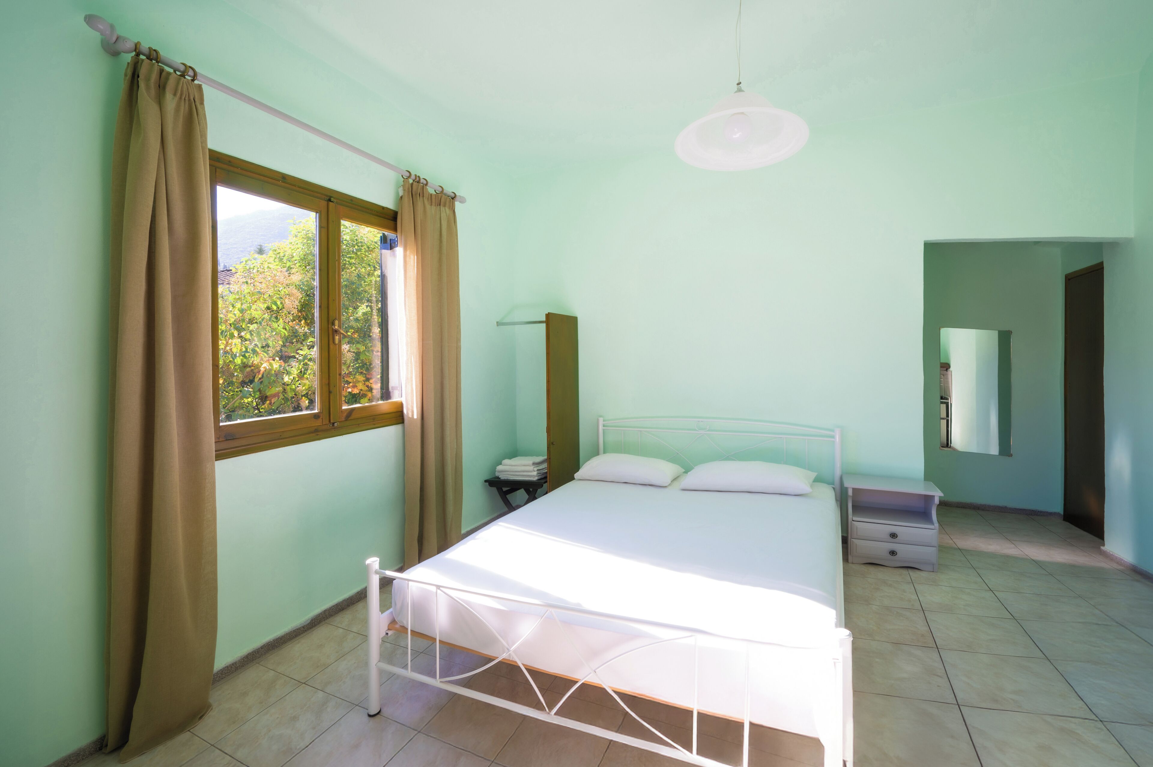 A bedroom with a bed, curtains, a bedside table, a lamp, and a window with a view of greenery.
