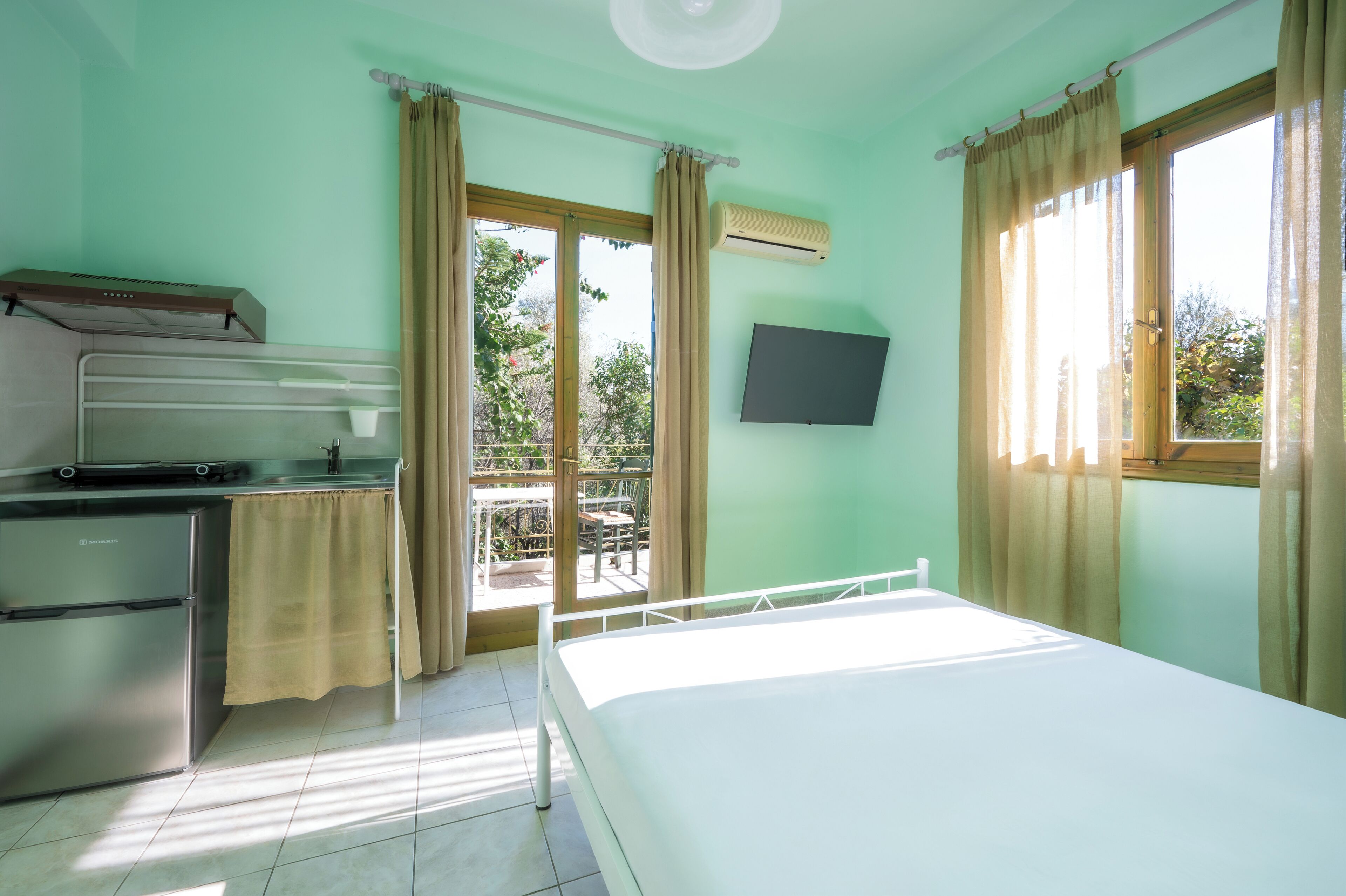 A room with a bed, a small kitchenette, a TV, and a balcony with curtains.