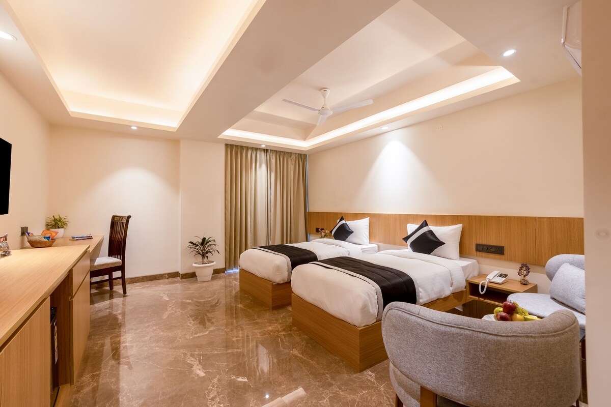 A hotel room with a desk, two beds, a chair, a small table with fruit, and a ceiling fan.