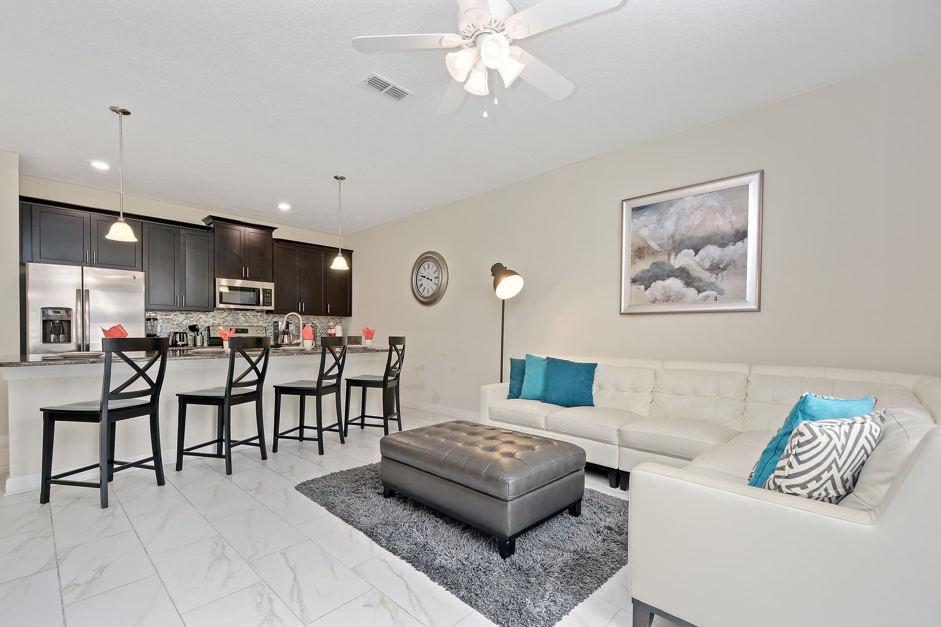 A modern kitchen with dark cabinets, a white countertop, and stainless steel appliances. There is a dining area with a white sofa and a gray ottoman.