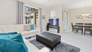 A living room with a white sofa, a dark brown ottoman, a TV on a stand, a dining table with chairs, and a chandelier. - Wonderful 4Bd With Pool at Storey Lake Resort 4819 (Kissimmee)