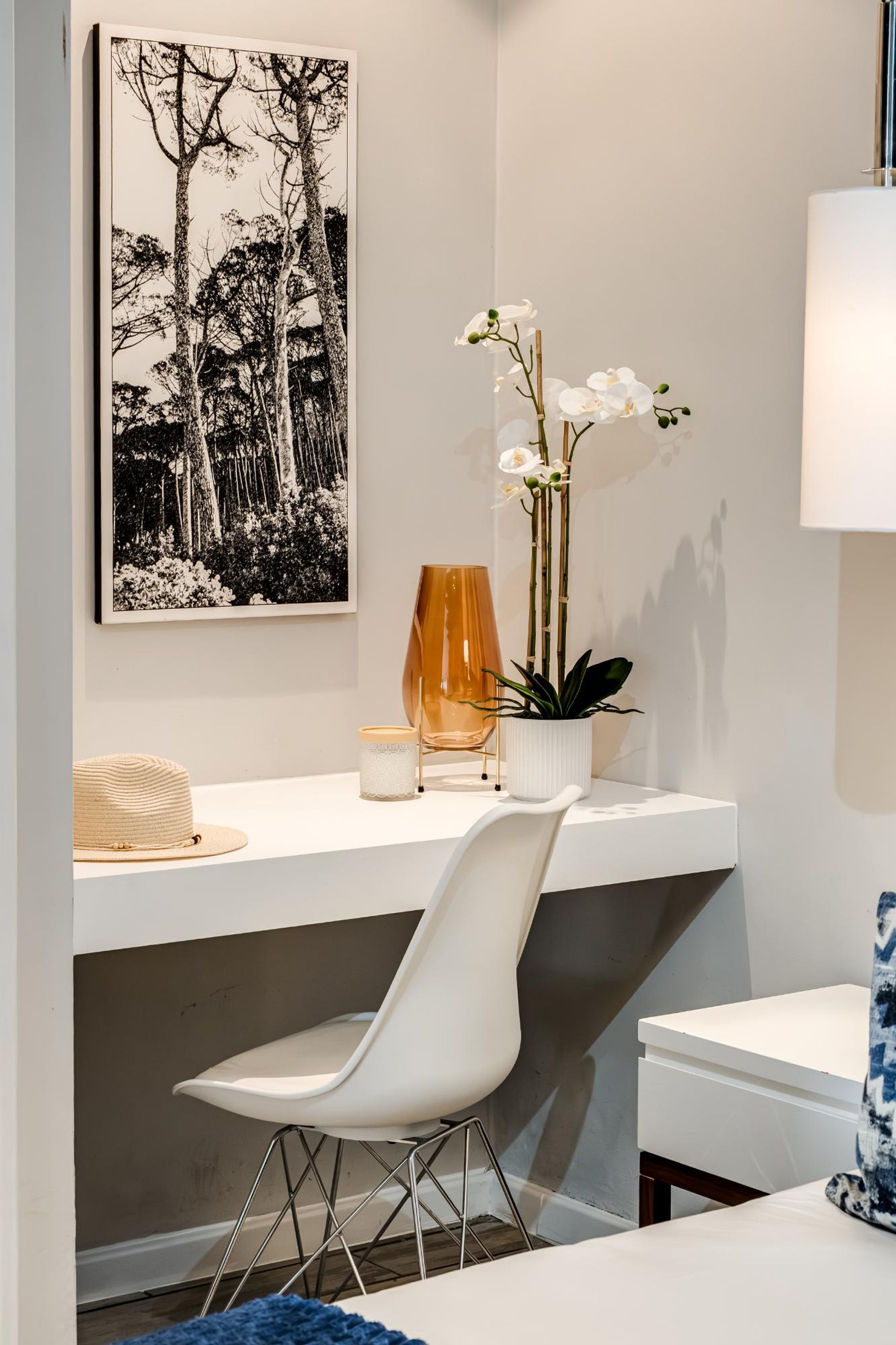 A modern room with a white desk, a white chair, a framed black and white photograph, a potted orchid, and a lamp.