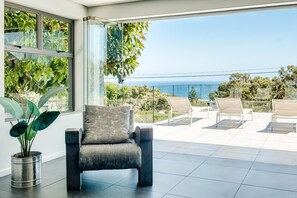 A modern living room with a large window overlooking a balcony with a view of the sea. - Ocean View Camps Bay 5bed Villa w Pool Valtameri (Cape Town)