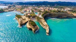 Vue aérienne d’une ville côtière, avec ses eaux turquoise limpides, ses formations rocheuses et un quartier résidentiel densément bâti, où l’on aperçoit des piscines et un accès à la plage.