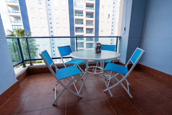 A balcony with a round table and blue chairs, overlooking a high-rise building.